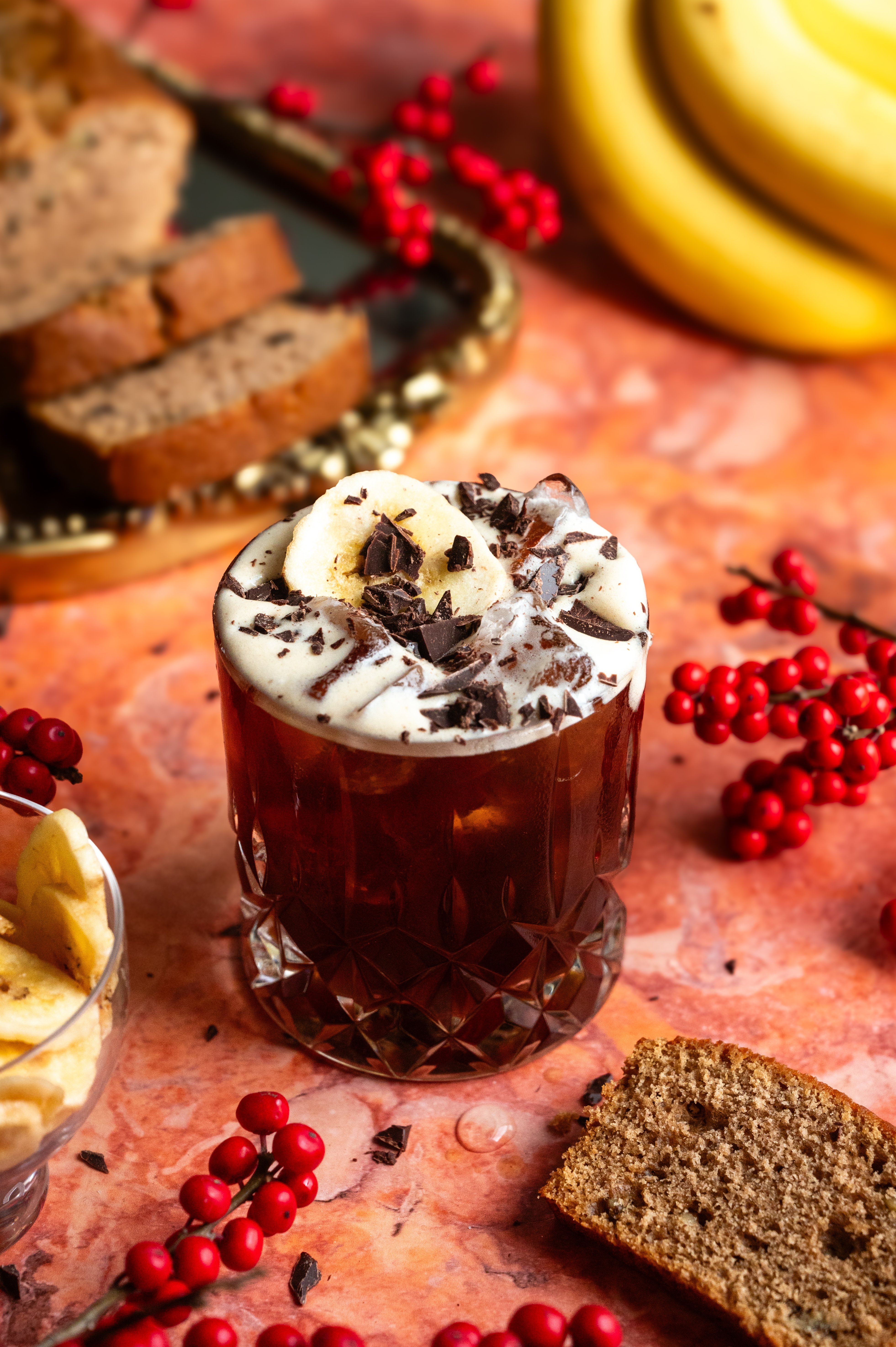 A rocks glass on an orange marble table filled with the banana bread carajillo and garnished with shaved chocolate and a banana chip.