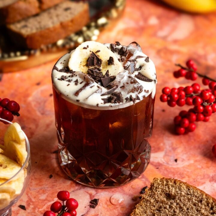 A rocks glass on an orange marble table filled with the banana bread carajillo and garnished with shaved chocolate and a banana chip.