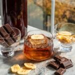 A banana bread old fashioned in a small rocks glass with the batched container, behind it, and surrounded by banana chips and chocolate. Its. also on a marble table in front. of a window.