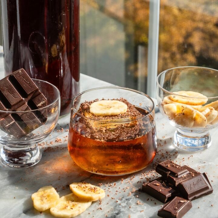A banana bread old fashioned in a small rocks glass with the batched container, behind it, and surrounded by banana chips and chocolate. Its. also on a marble table in front. of a window.