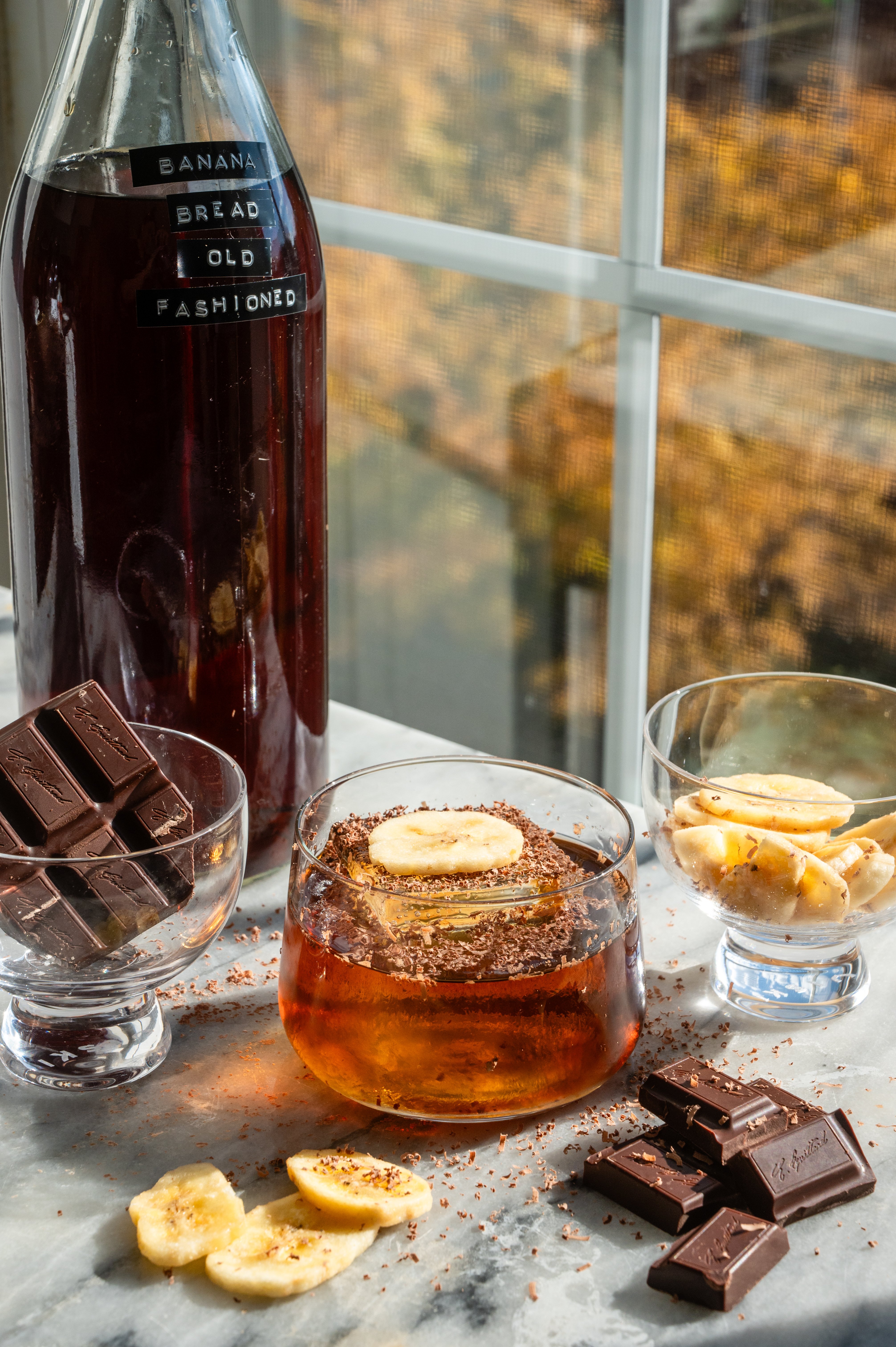 A banana bread old fashioned in a small rocks glass with the batched container, behind it, and surrounded by banana chips and chocolate. Its. also on a marble table in front. of a window.