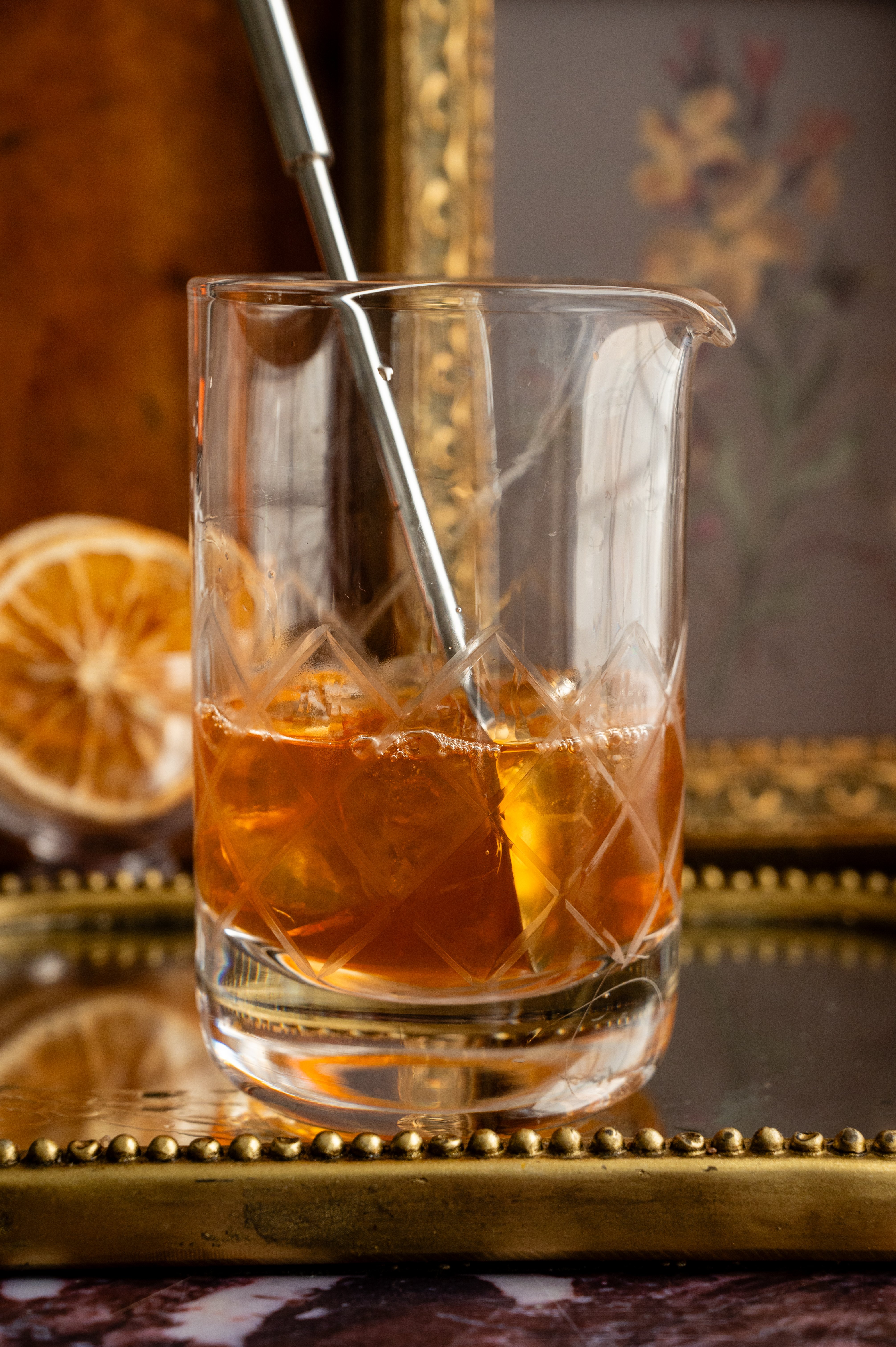 A mixing glass filled with ice and the old fashioned ingredients. A spoon is stirring the cocktail.