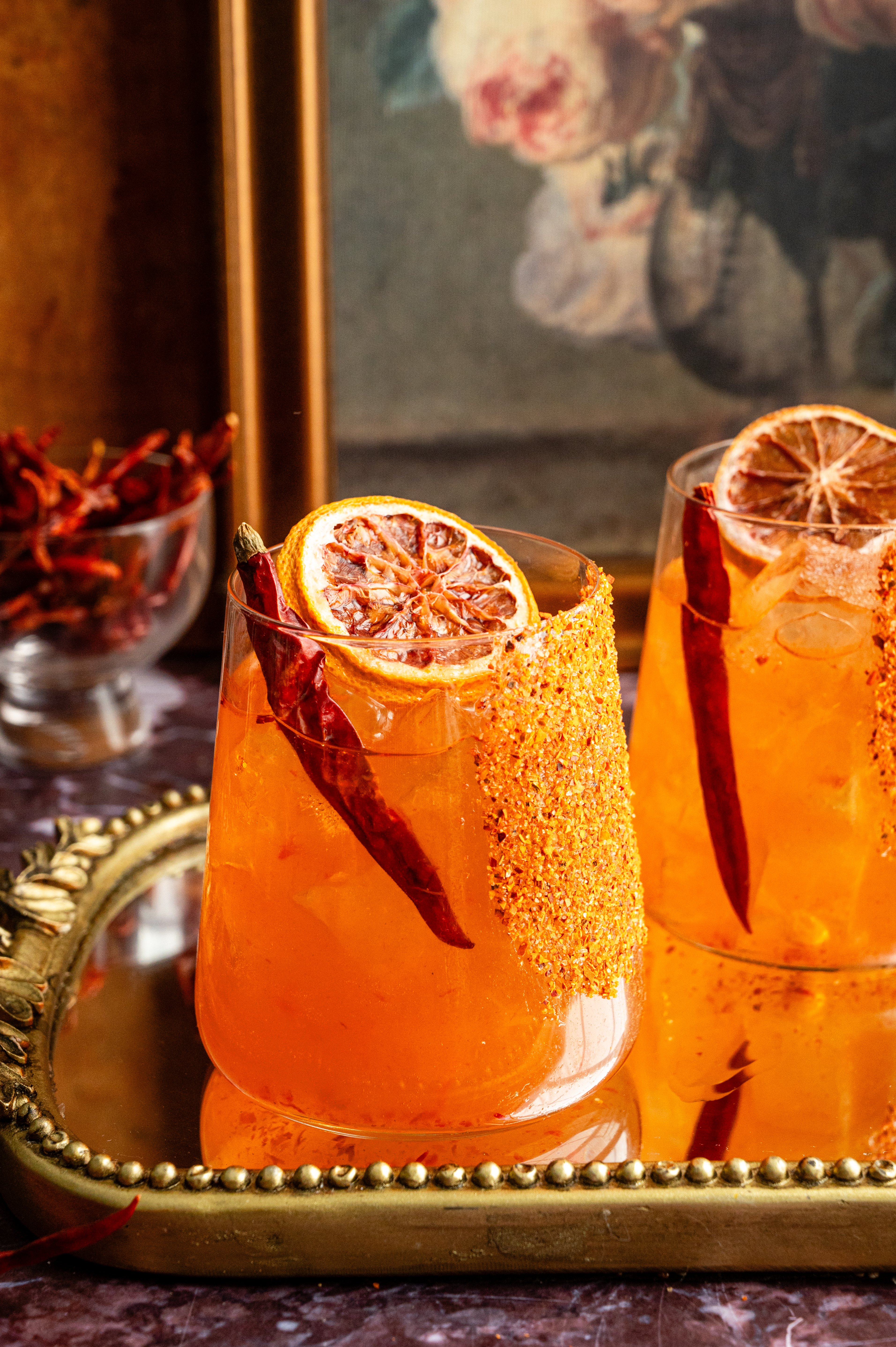 Calabrian chili margaritas on a mirrored tray and. garnished with a dried chili pepper and. dehydrated orange. 