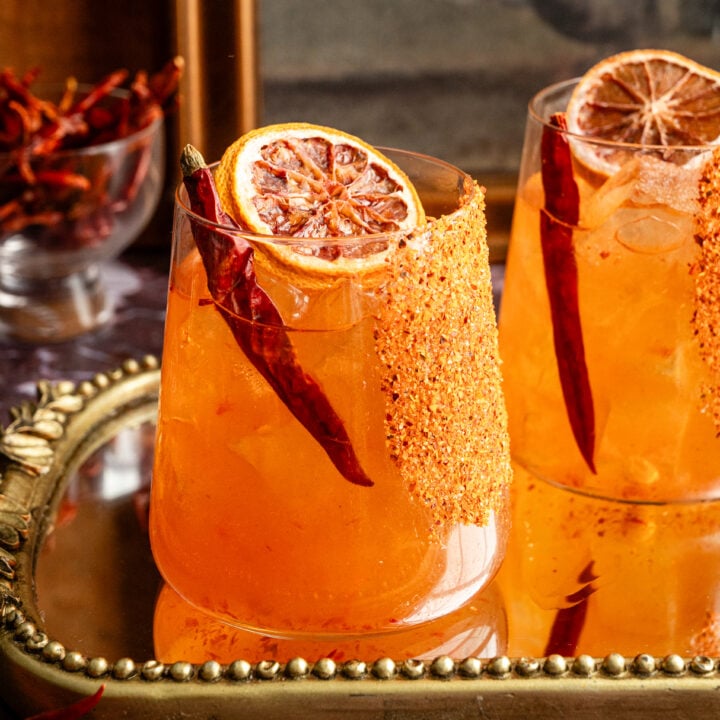 Calabrian chili margaritas on a mirrored tray and. garnished with a dried chili pepper and. dehydrated orange.