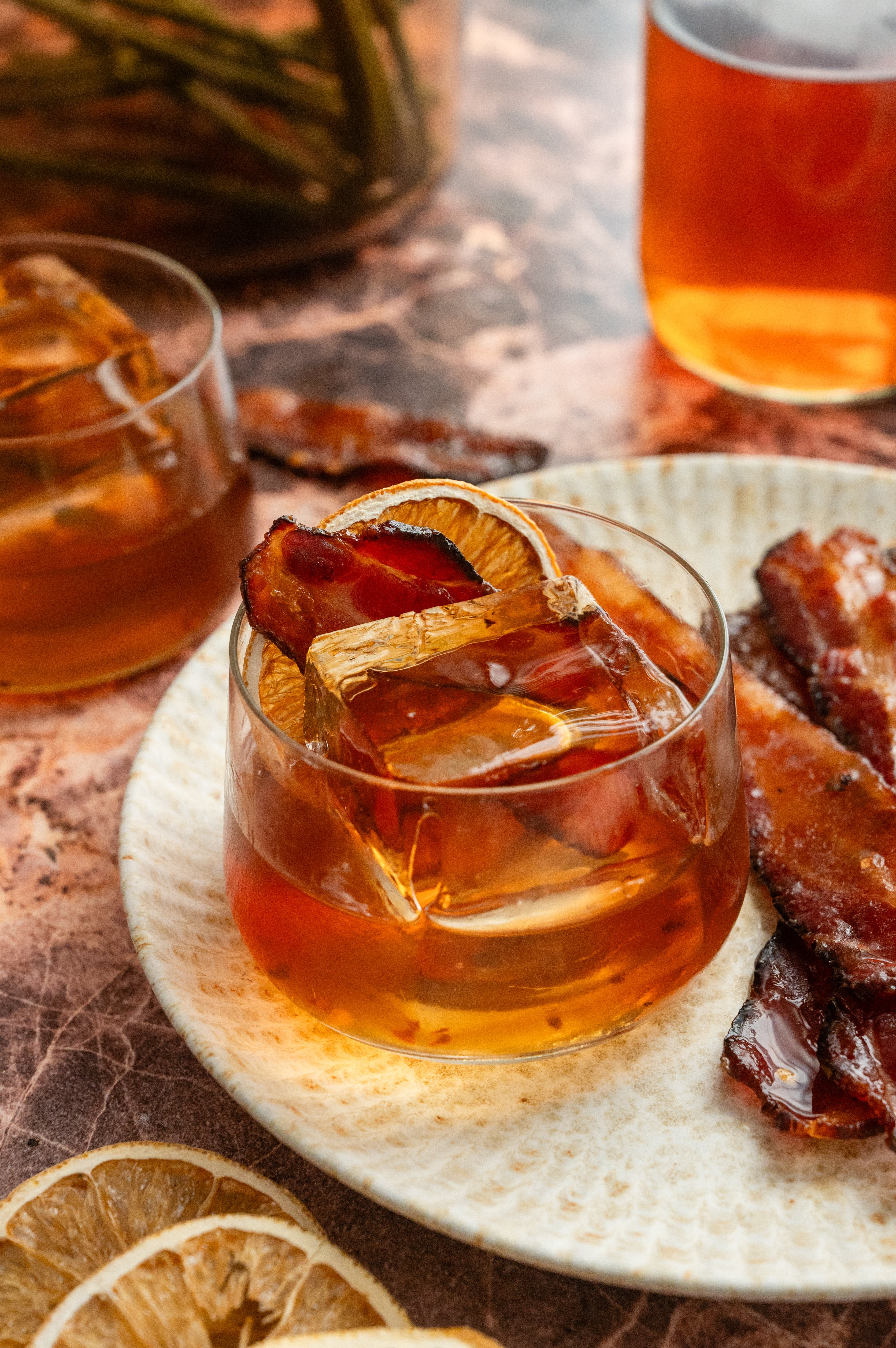 The finished cocktail in a low rocks glass with a large ice cube and garnished with a dehydrated orange and a slice of millionaire bacon.