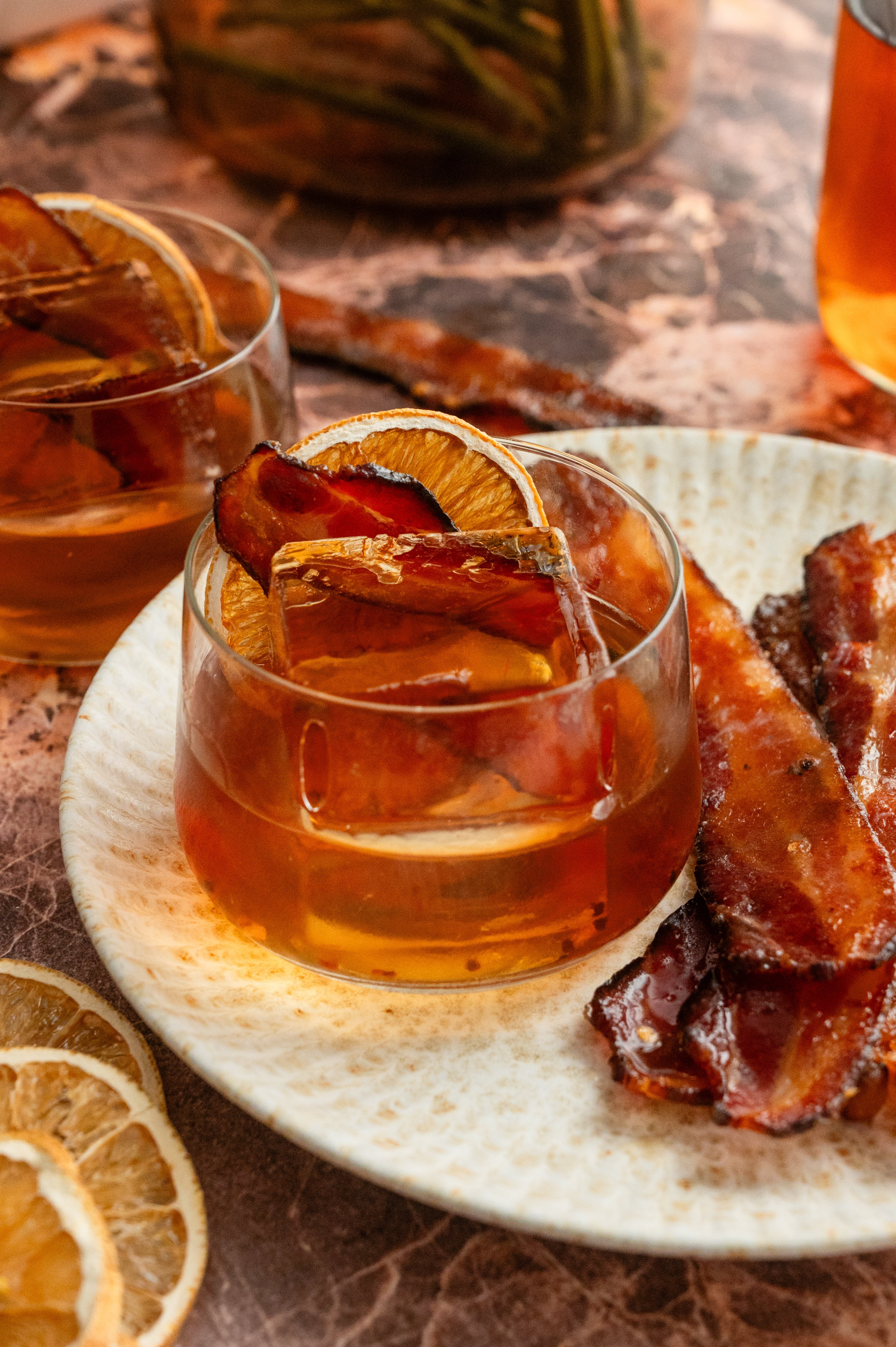 The finished cocktail in a low rocks glass with a large ice cube and garnished with a dehydrated orange and a slice of millionaire bacon
