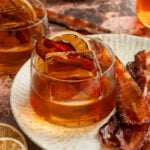 The finished cocktail in a low rocks glass with a large ice cube and garnished with a dehydrated orange and a slice of millionaire bacon