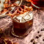 The shaken maple bacon bourbon espresso old fashioned in a wavy rocks glass and garnished with bacon.