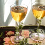 two elderflower manhattans in nick and. Nora glasses backlit on a floral tray.