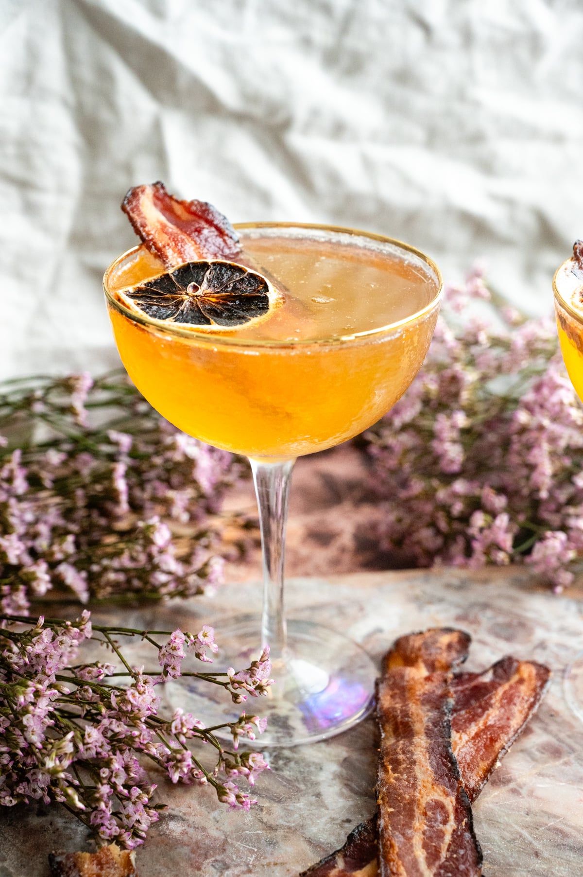 The cocktail in a coupe glass garnished with millionaire bacon and lemon. On a pink marble table.