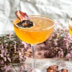 The cocktail in a coupe glass garnished with millionaire bacon and lemon. On a pink marble table.