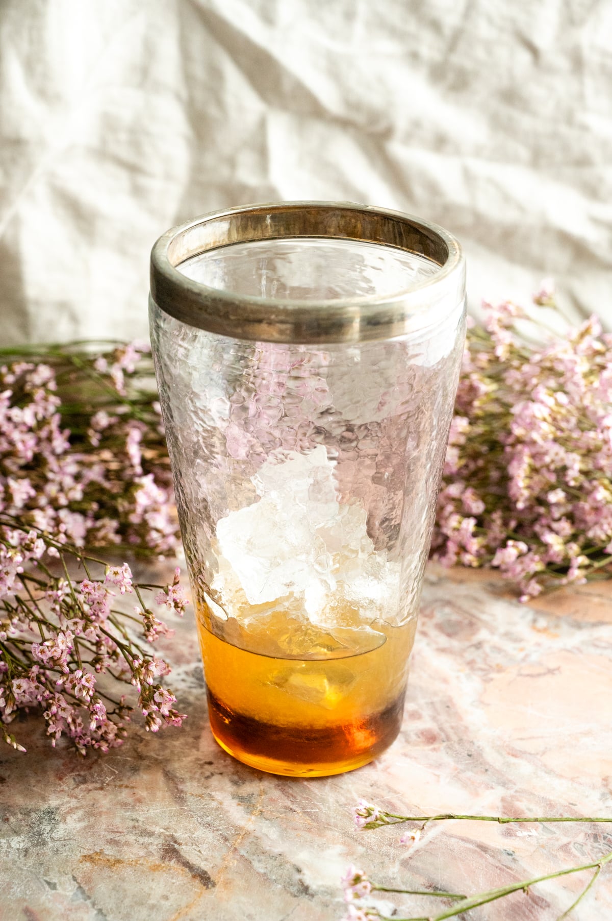 A glass shaker filled with the cocktail ingredients and ice on a pink marble table.