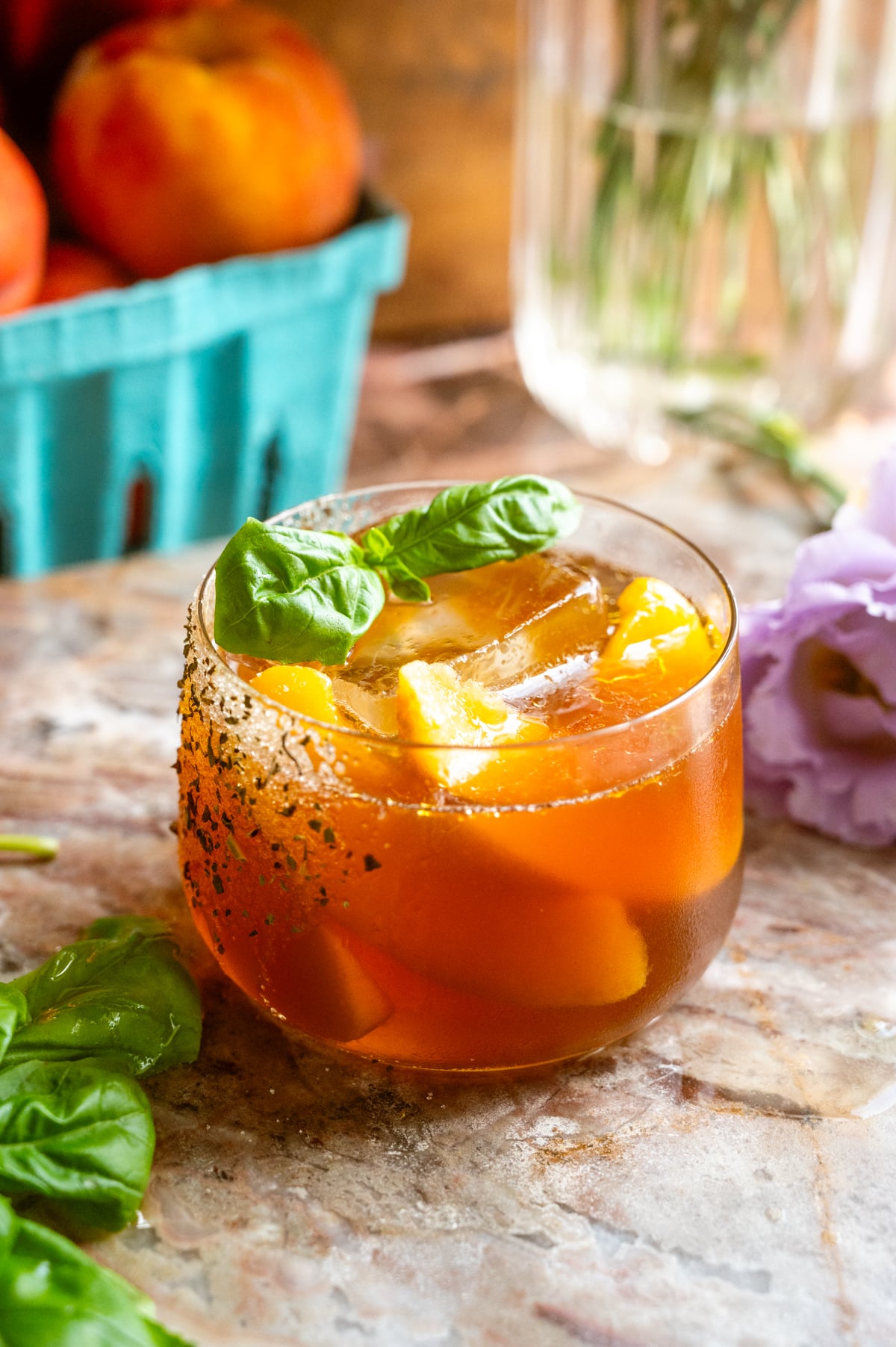 A short rocks glass filled with the cocktail and garnished with frozen peaches and couple of basil leaves on a pink marble slab.