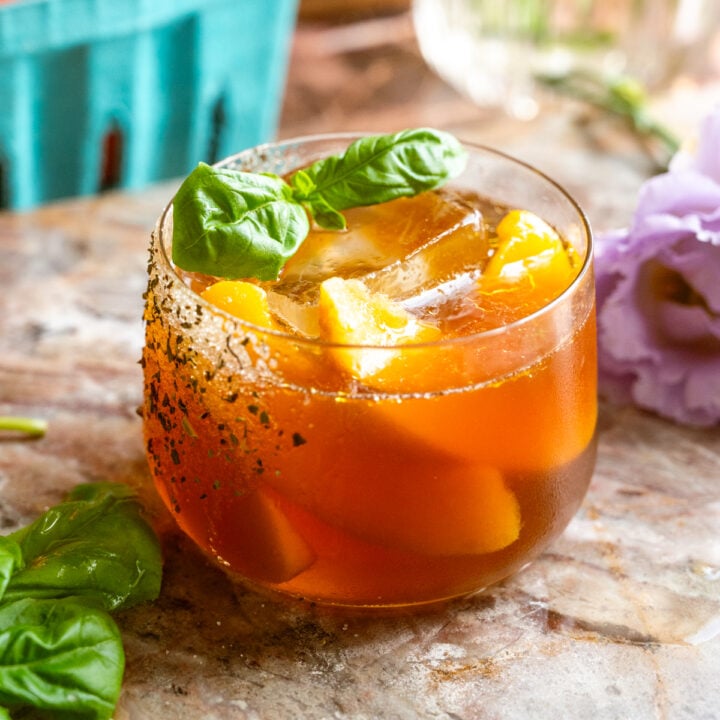 A short rocks glass filled with the cocktail and garnished with frozen peaches and couple of basil leaves on a pink marble slab.