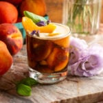 Peach carajillo served over frozen peaches in an etched glass and topped with a peach slice. Its placed on a pink marble slab and has purple flowers next to it.