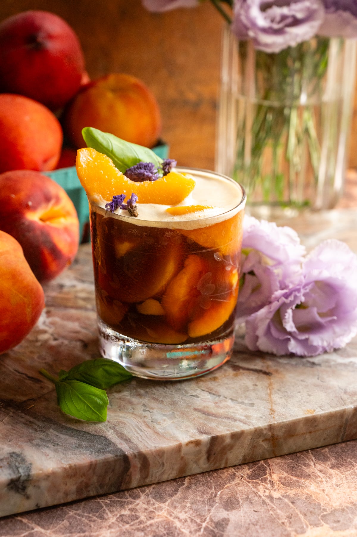 Peach carajillo served over frozen peaches in an etched glass and topped with a peach slice. Its placed on a pink marble slab and has purple flowers next to it.